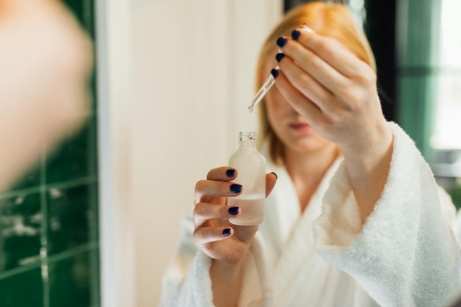 Woman applying vitamin C serum in front of a bathroom mirror showing the morning routine step